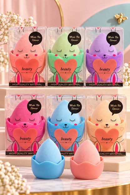 Six boxed makeup sponge colors with plain blue and pink sponge-and-stand sets in front; one selected boxed set per purchase